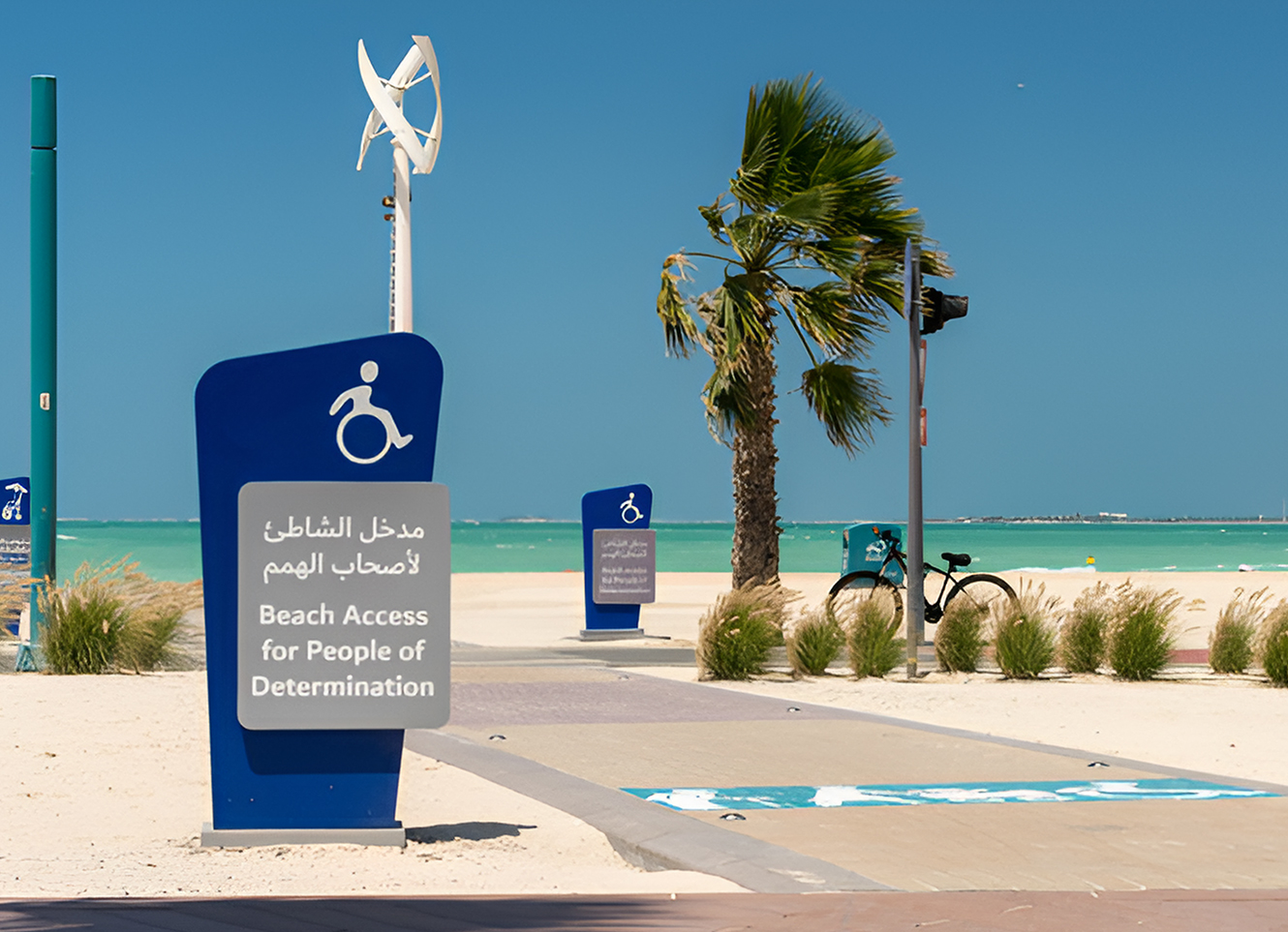 A beach access sign for People of Determination near a pathway leading to the shore, with turquoise water, palm trees, and accessible beach markings.