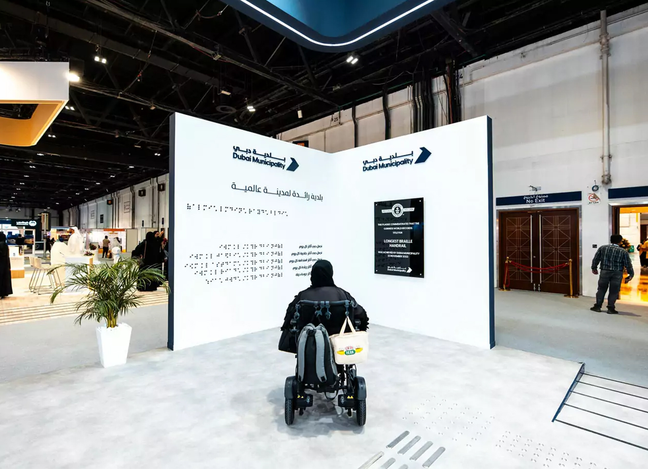A visitor using a wheelchair interacting with a Dubai Municipality accessibility exhibit featuring Braille text and an information wall at an indoor event.