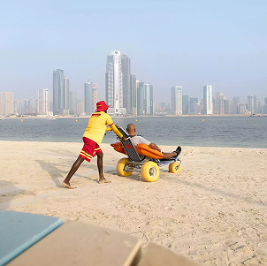 Lifeguard pushing a person in a beach wheelchair along the shoreline with city skyline in the background