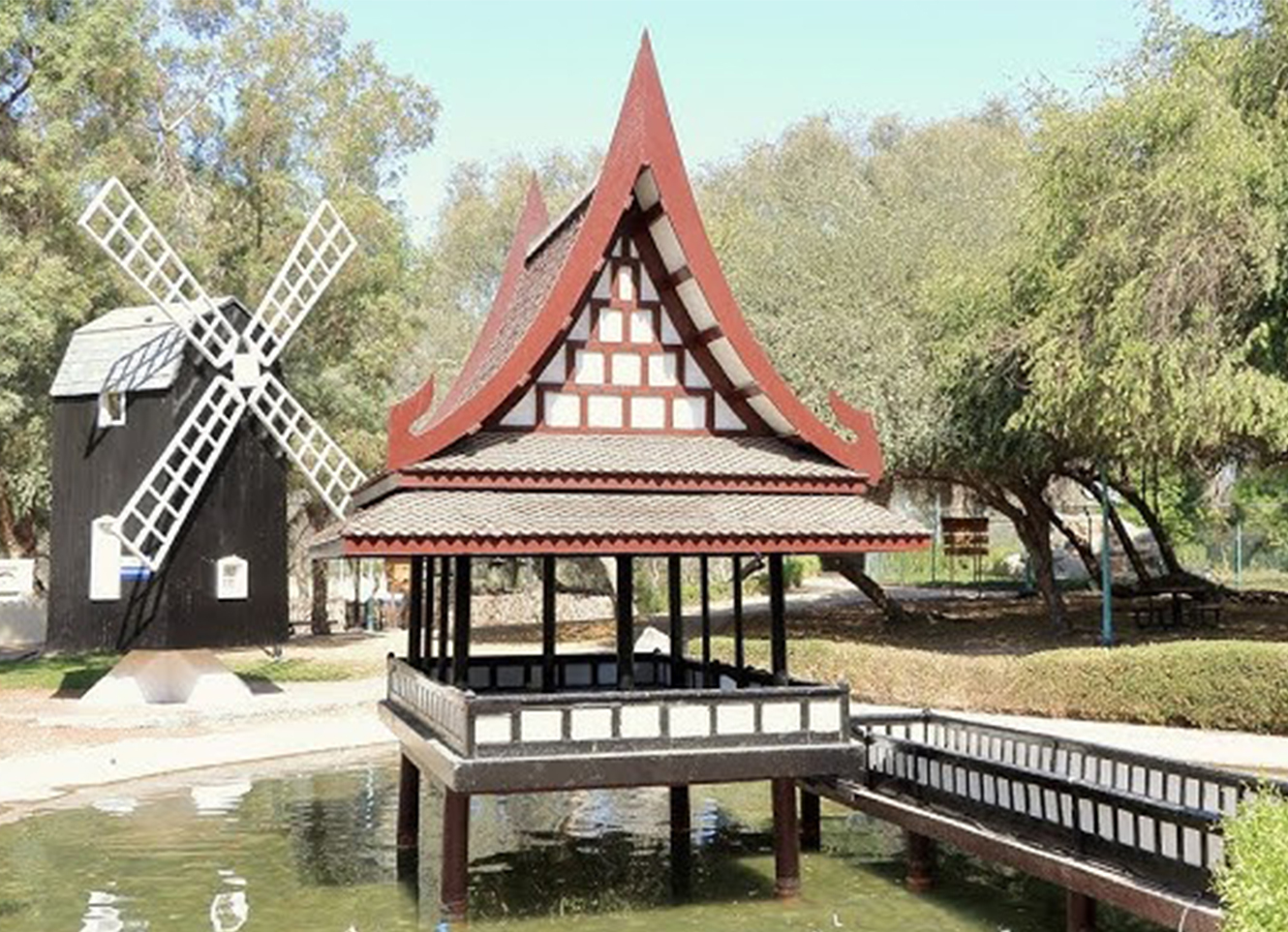 A miniature cultural-style pavilion on a pond with a traditional windmill structure in the background, surrounded by trees in a public park.